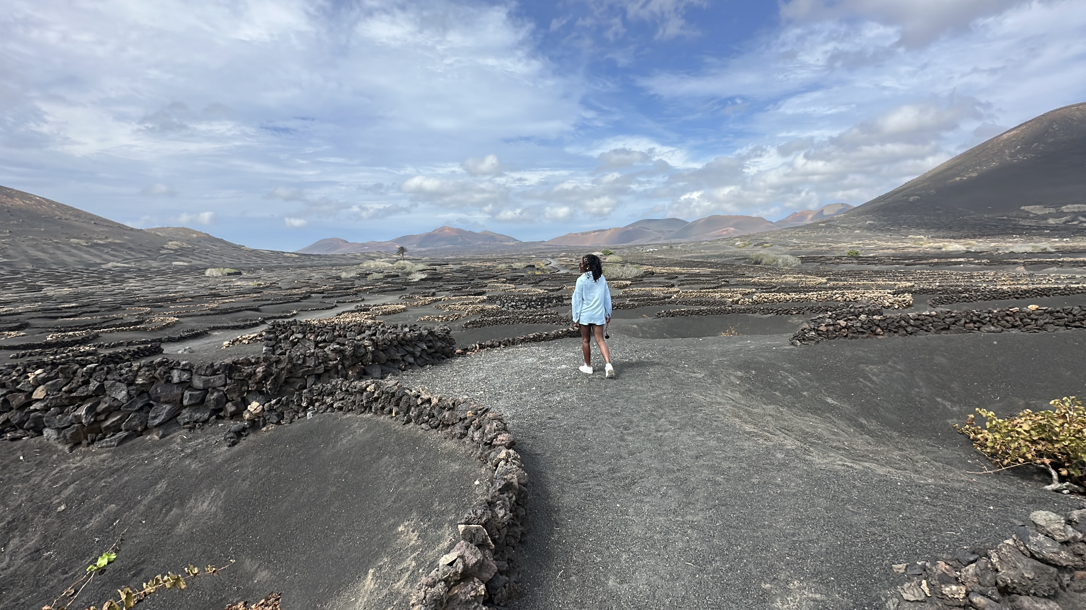 Neola Husbands in Lanzarote, Canary Islands — tiny figure standing in a black volcanic vineyard (La Geria), mountains in the distance, surface that looks like Mars.