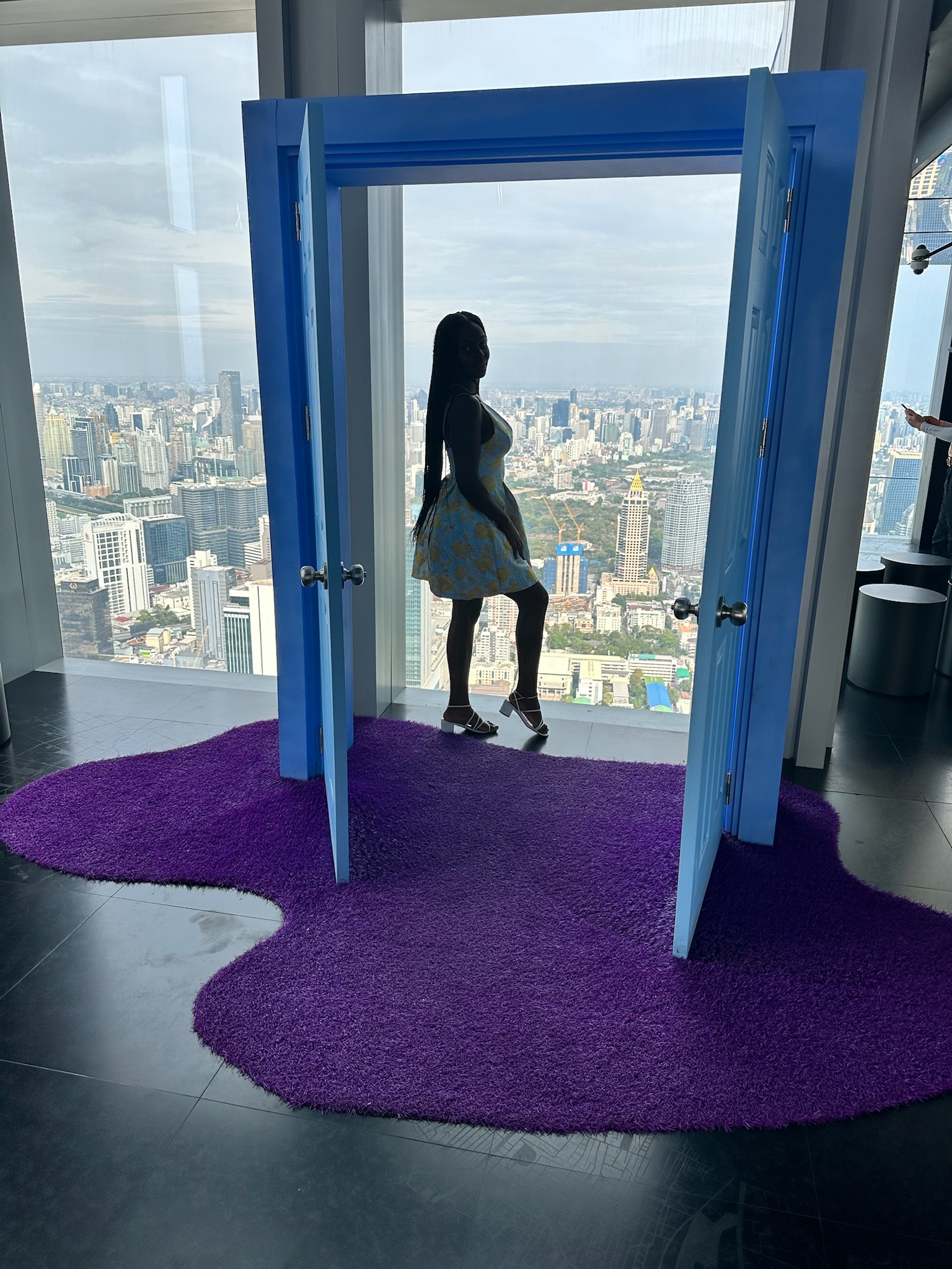 Bangkok — Neola stepping through blue doors onto a purple shag rug, city skyline behind.